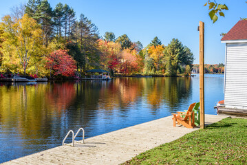 Deserted wooden riverside boardwalk on a sunny autumn day. Two colourful adirondack chairs are on...