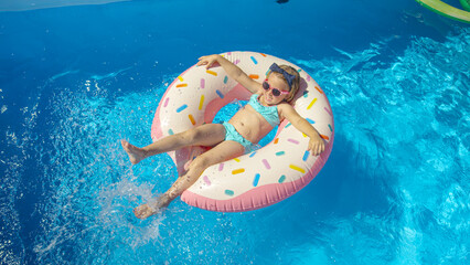 TOP DOWN: Happy little girl splashing water on a floatie donut in swimming pool