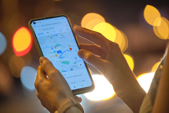 Close up of female hands holding cellphone with Google maps navigation app searchig for direction with blurred city street lights on background at night. Kyiv, Ukraine - November 2, 2021.