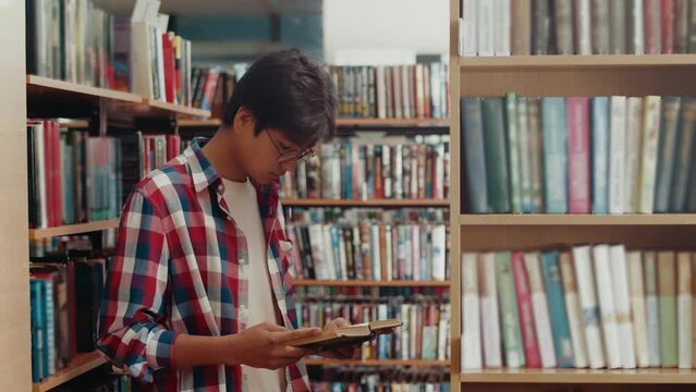 An Asian student in a shirt and glasses reads a book in library and then looks at the camera. An oriental man works the library