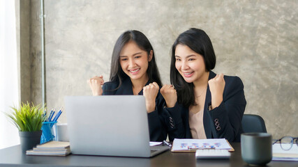 Two young Asian businesswomen show joyful expression of success at work smiling happily with a laptop computer in a modern office.