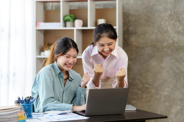 Two young Asian businesswomen show joyful expression of success at work smiling happily with a laptop computer in a modern office.