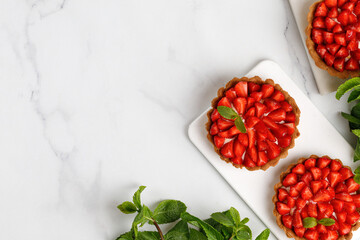 Tarts with strawberries and whipped cream decorated with mint leaves on white board and marble background. Top view, copy space.