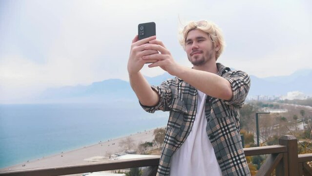 Young stylish blonde man takes selfie on the background of the sea