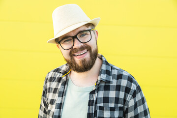 Portrait of positive young hipster man smiling over yellow wall store outdoor background - Handsome trendy bearded guy with hat and glasses standing outdoors - Fashion and trendy youth people concept