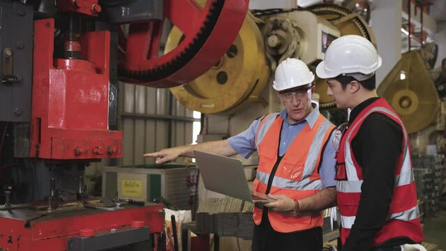 Senior engineer wear safety goggles and helmet with holding and using laptop to coaching young engineer to operate hydraulic pump machinery in factory. Two industry maintenance workers in warehouse. 