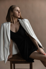 beautiful caucasian woman in a black dress and white jacket sits on a chair in the studio on a beige background