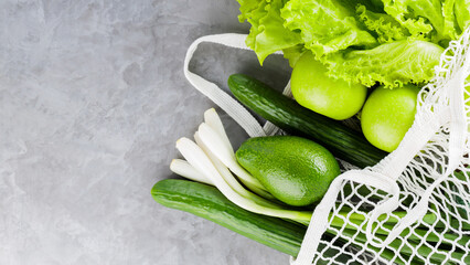 Reusable grocery net bag with fresh vegetables and fruits on gray concrete. Zero waste mesh bag with green fruits and vegetables on a gray background. Eco-friendly shopping. Copy space