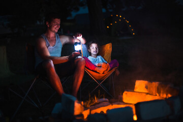 Fototapeta na wymiar Dad and daughter sit at night by the fire in the open air in the summer in nature. Family camping trip, gatherings around the campfire. Father's Day, barbecue. Camping lantern and tent