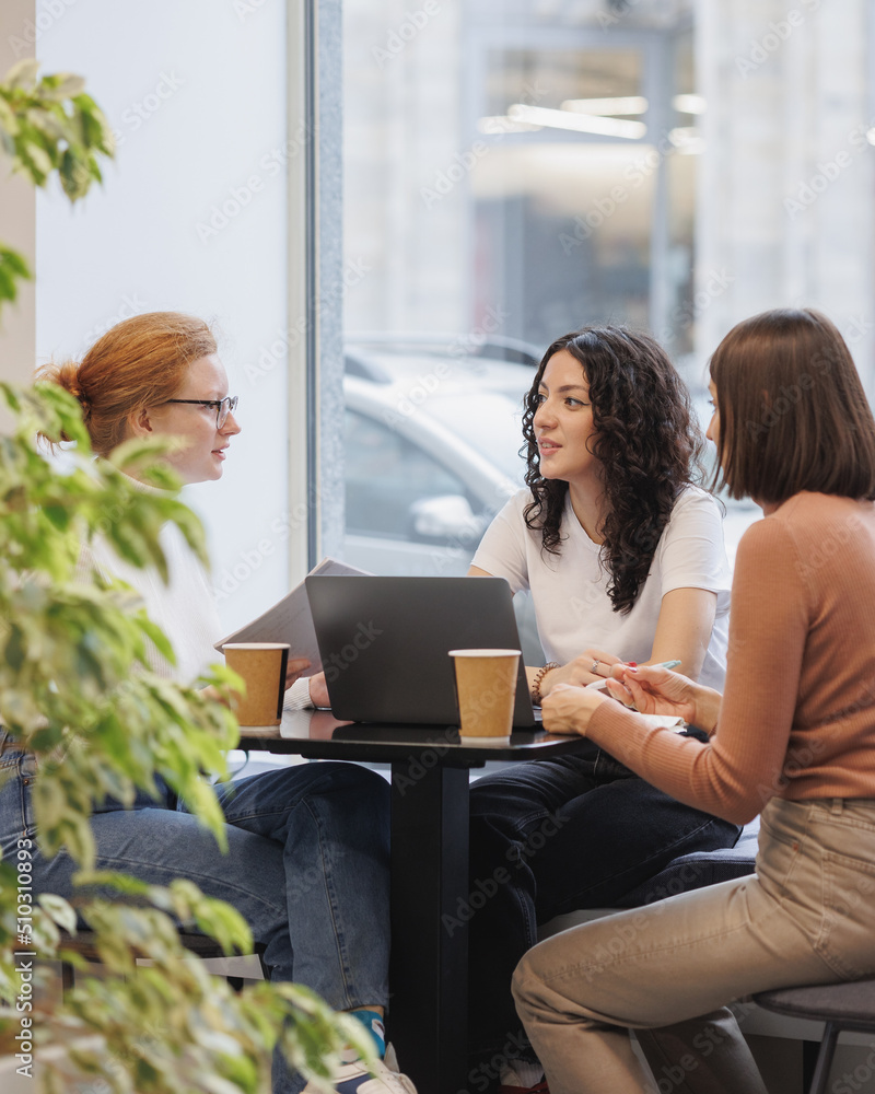 Wall mural three female colleagues or students are working on a laptop and discussing a project or creative term paper. three female friends in coworking work online in an intranet or video chat for negotiations