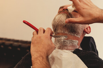Shaving with a straight razor in a barbershop. A bearded old man being shaved in a barbershop....