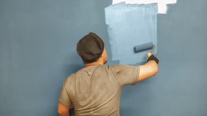 A rear view of an artist in a gray T-shirt, black cap and gloves, who is painting a wall with a paint roller.