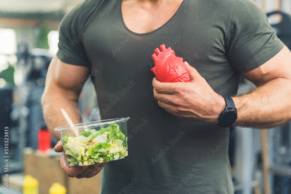 Wall mural Unrecognizable muscular fit caucasian guy in a t-shirt holding a bowl of green vegetable salad and a fake human heart. High quality photo