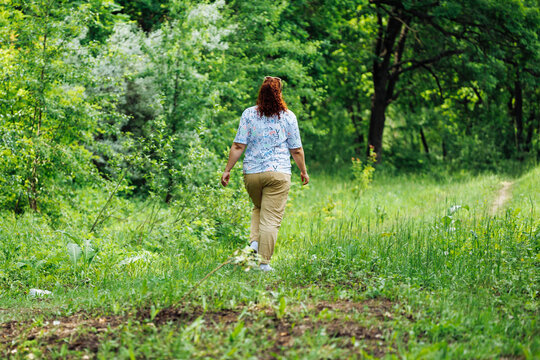 Back view of young buxom woman with long curly red hair in ponytail, wearing blue blouse with floral pattern, beige trousers, walking on path in park forest among green trees. Summer, nature, relax.