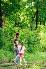 Back view of family walking in park forest around green trees, talking, having fun. Little cheerful daughter holding hand of middle-aged bearded man father. Summer activities, travelling. Vertical.