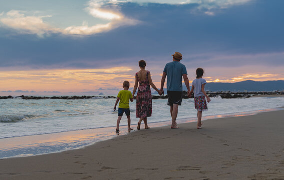A family of four strolls along the beach at sunset. Golden hues reflect on the ocean. Waves gently crash, laughter fills the air, creating cherished moments by the sea.