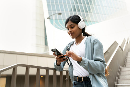 Young Beautiful Indian Girl Using Mobile Phone Communication Online, Reading Text Message Walking On The Street. Asian Hipster Wearing Wireless Headphones Listening Music Outdoors. Modern Technology 