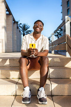 Happy, young African American man using mobile phone outdoors sitting on stairs. Vertical image.