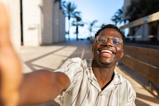 Happy young black man with glasses taking selfie looking at camera.