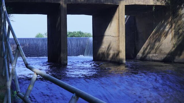 Camera zoom in forward. Shade and view through channel of watercourse under the bridge. Old concrete dam with water flowing rapids. During the rainy season in rural Thailand.