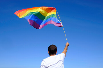 Adult man in white t-shirt waves rainbow flag with blue sky and ocean in the background.