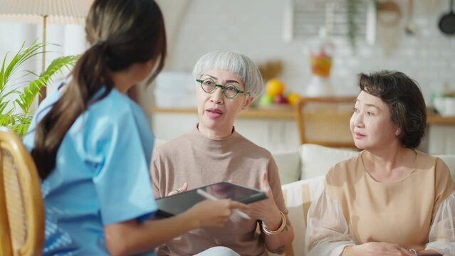 happiness Cheerful elderly woman and men talking with female caregiver nurse doctor having health checking consult at living area,Caretakers with senior couple sitting in living room at nursing home 