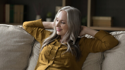 Obraz premium Pretty middle-aged woman with long grey hair sit on cozy sofa smile enjoy relaxation in modern house, fresh conditioned air and free time alone at home. Repose, natural beauty of older female concept