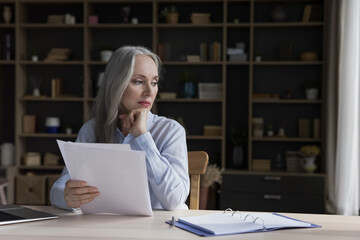 Attractive brooding aged businesswoman sit at workplace desk holds papers feels exhausted...