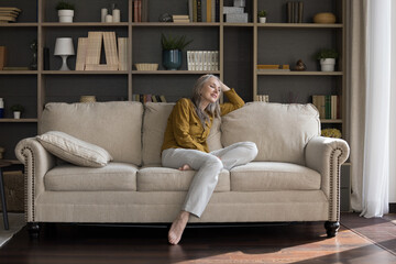 Obraz premium In cozy light living room middle-aged serene beautiful woman sit on sofa looks pensive enjoy free time, daydreaming relaxing alone at home. Homeowner person, tenancy, modern furniture store ad concept