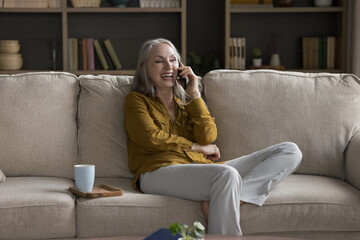 Obraz premium Happy attractive senior woman laugh feels overjoyed enjoy pleasant conversation on cellphone relax on couch alone at home. Older generation using modern wireless tech, communication, good news concept