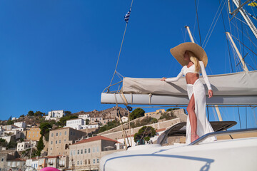 Beautiful young blond woman standing on catamaran at sunny summer day at Hydra marina bay, Greece
