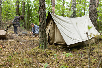 Boys building a fire in a fire pit made out of bucket near the tent in the forest. Starting a campfire- Starting a fire using a fire striker- bushcraft and primitive skills.