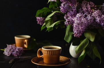 Obraz na płótnie Canvas evening tea with friend in cozy setting, Brown mugs in blur. Dark key. soft focus on lilac flowers.