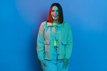 Portrait of dark haired young Caucasian delighted woman wearing stylish jacket posing isolated on neon light background, looking at camera with amazed facial expression.