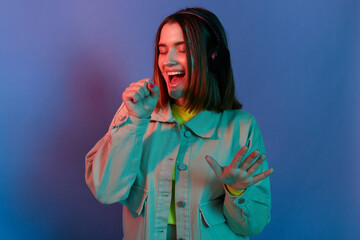 Horizontal shot of beautiful dark haired woman wearing stylish jacket posing isolated on neon light background, listening music, keeping hand near mouth, imagines she is singing on microphone.