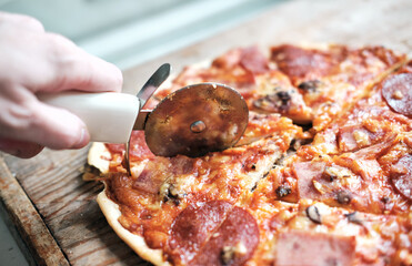the hand holds a round knife and cuts a pizza with salami, close-up. round pizza with tomatoes and salami on a wooden background.Italian fast food.
