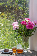 Delicious hot tea on the windowsill at home at summer day near garden and beautiful bouquet of flowers pink peonies, close up. Hot black tea in a glass teapot and cup