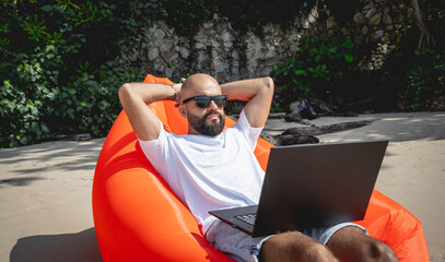 Young man working on his laptop on the beach while sitting on an inflatable sofa