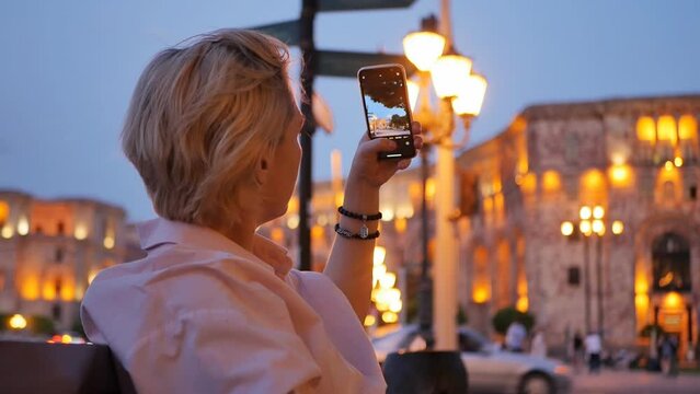attractive caucasoid blonde sitting on a bench takes pictures on a smartphone of beautiful city buildings in the center of a big city illuminated by evening lanterns.