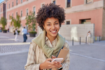 Cheerful female model with curly hair checks email on smartphone carries notepads dressed in casual clothes expresses positive emotions strolls outdoors against urban background uses cellular app