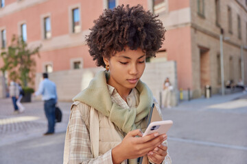 Fototapeta na wymiar Curly haired millennial girl focused at smartphone scrolls news in internet wears shirt and jumper tied over shoulders poses outdoors in urban setting blurred background. Modern technologies