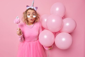 Obraz na płótnie Canvas Horizontal shot of amazed shocked young woman wears festive dress unicorn headband and dress stares at camera celebrates special occasion holds bunch of balloons isolated over pink background