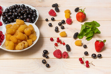 Berries of yellow raspberries and black currants in a white bowl. Berries scattered on a wooden table