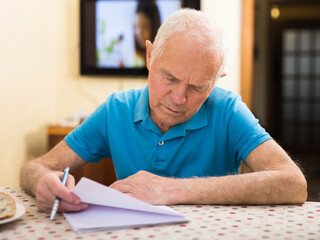 Elderly man writes a letter on a sheet of paper while sitting at a table in room