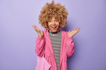 Happy amazed woman with curly hair spreads palms has surprised excitd expression wears casual striped jumper pink jacket and fabric bag isolated over purple background. Human reactions concept