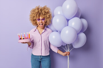 Festive occasion and celebration concept. Emotional woman with curly hair holds sweet cake and bunch of helium balloons exclaims loudly wears shirt jeans and sunglasses isolated over purple wall