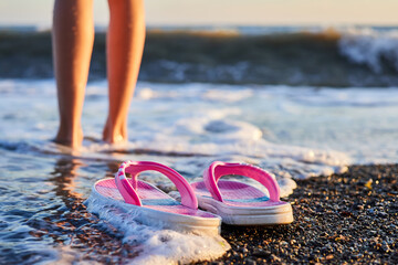 Pink children's slippers on a pebble beach with girl legs on the background