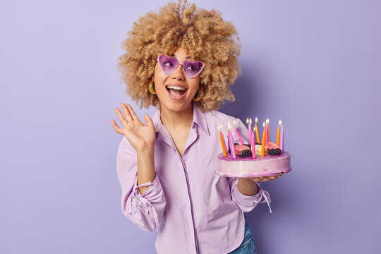 Waist up shot of happy birthday girl with curly blonde hair wears trendy heart shaped sunglasses and shirt holds delicious cake with burning candles isolated over purple background. Festive event
