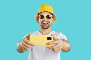 Cheerful carefree guy making selfie shot on mobile phone on light blue background. Portrait of smiling young man in panama and sunglasses with yellow cellphone making selfie or video recording.