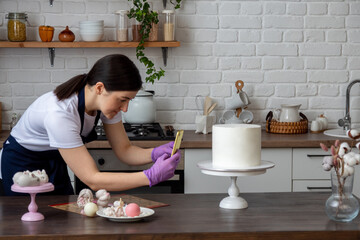 Woman in an apron holds smartphone in her hands and takes pictures of the cake. There are chocolate figurines on the table. Selective focus. Photos about confectioners, food, hobbies.